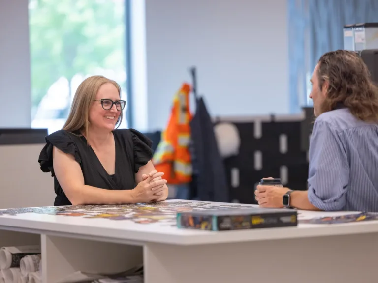 Two MacKay colleagues talk at an office table; one holds a coffee cup, with a high‑visibility vest and hard hat blurred in the background.