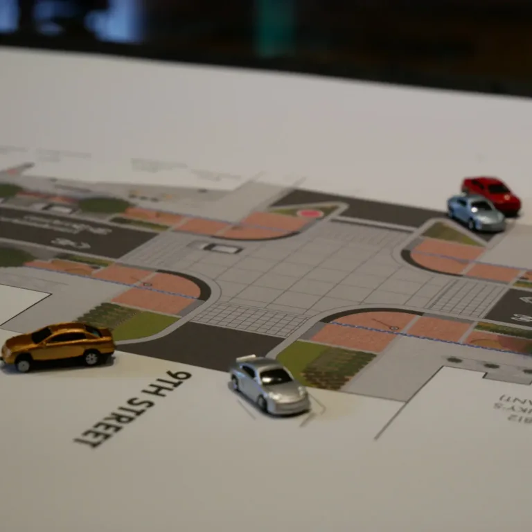 Small toy cars are placed on a printed urban design plan showing an intersection on 9th Street with crosswalks, sidewalks, and landscaped areas.