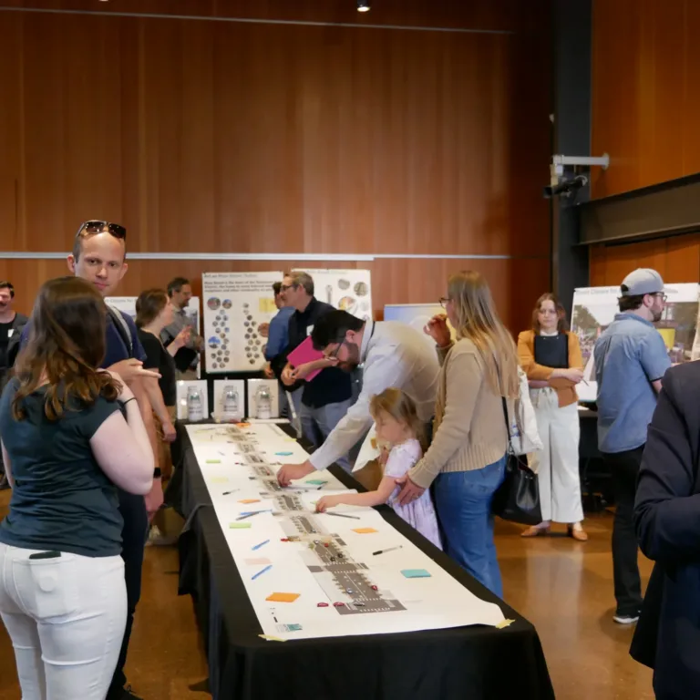 Attendees at an indoor community workshop examine a long street plan on a table covered with sticky notes while others discuss posters around the wood‑paneled room.