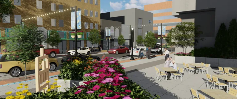 Rendering of a downtown street with bright flower planters, outdoor café seating, string lights, cars and bikes, and “Vancouver” banners among modern buildings on a sunny day.