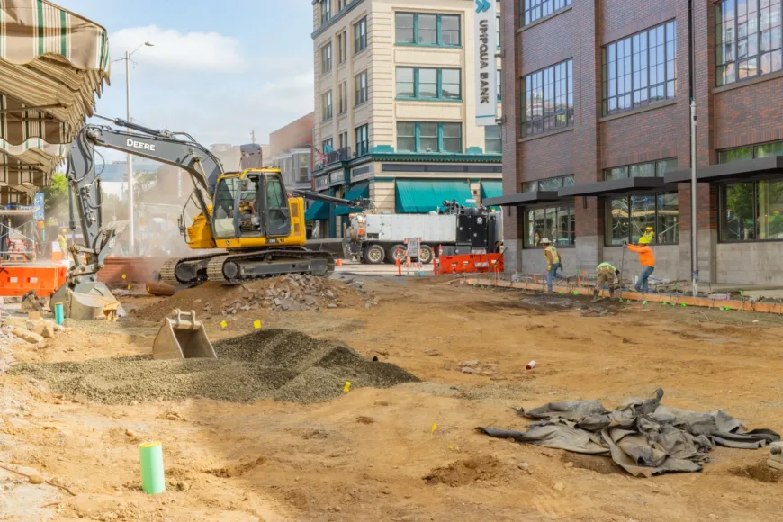 Downtown street construction scene with a yellow excavator and crew forming a sidewalk beside brick buildings, surrounded by orange barriers, cones, and piles of dirt and gravel.