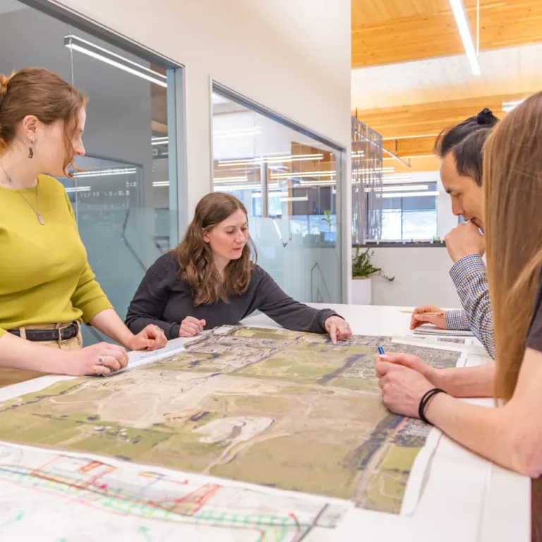 MacKay team collaborating around a conference table, reviewing large aerial site maps and plan drawings in a modern glass-walled office.