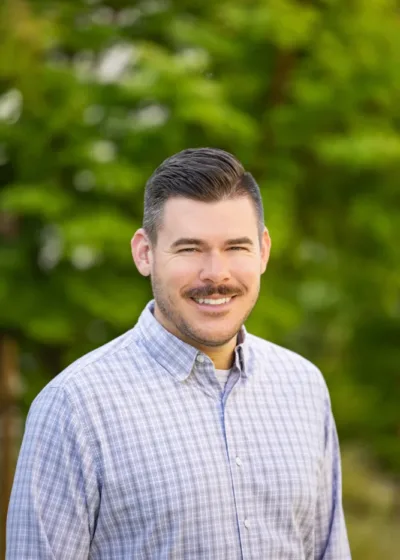Headshot of Michael Strachan smiling, with short dark hair and a mustache, wearing a light plaid button-down shirt against a blurred green outdoor background.