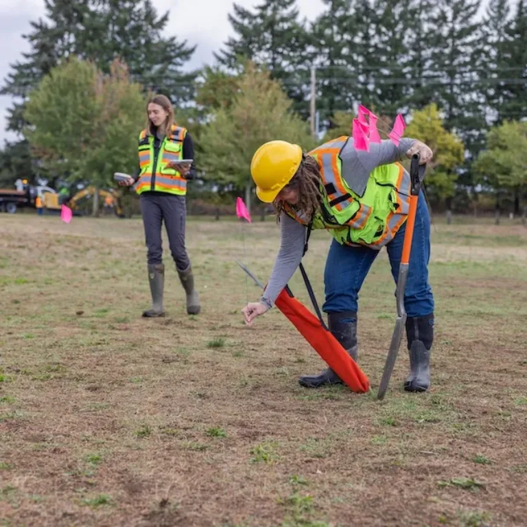 Utility workers in safety vests plant a pink flag in a field while another worker stands nearby