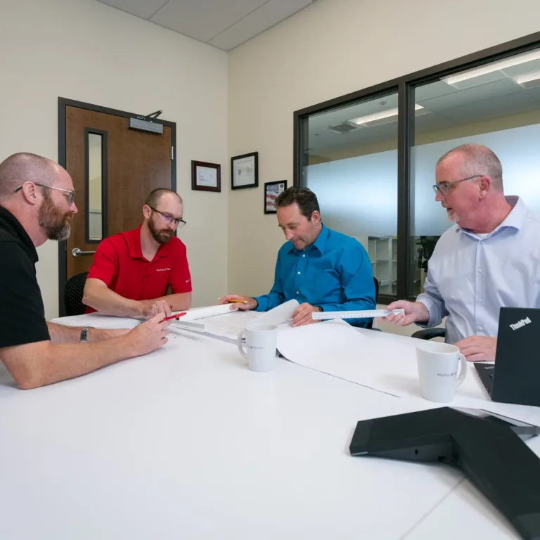 MacKay team members collaborate in a conference room, reviewing large engineering plans with drafting scales, mugs, and a laptop on the table.