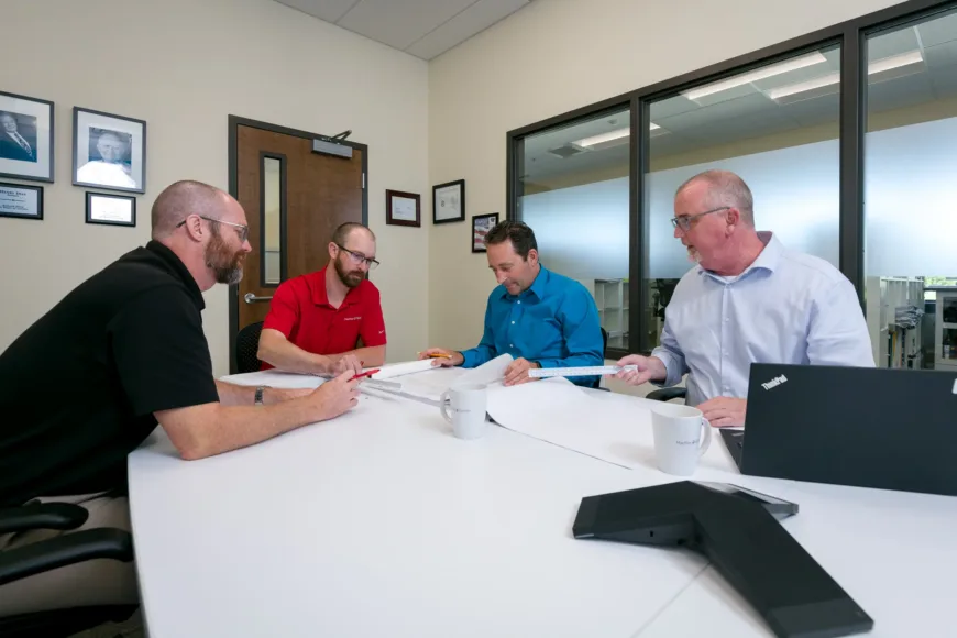 MacKay team members collaborate in a conference room, reviewing large engineering plans with drafting scales, mugs, and a laptop on the table.
