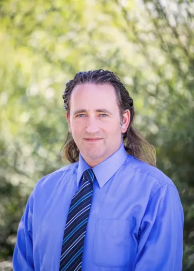 Headshot of Paul Harmsen wearing a light blue dress shirt and blue-striped tie, with shoulder-length hair and a slight smile against a blurred green background.
