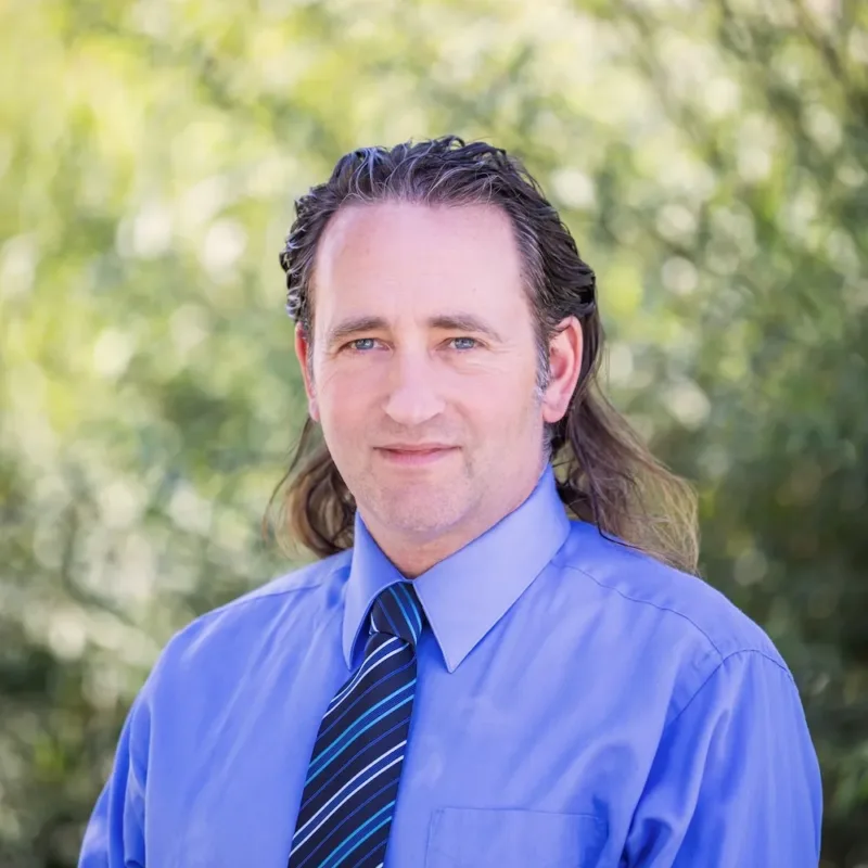 Headshot of Paul Harmsen wearing a light blue dress shirt and blue-striped tie, with shoulder-length hair and a slight smile against a blurred green background.