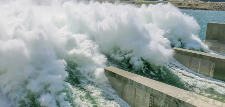 Massive ocean waves crash into concrete breakwater channels, sending white spray high into the air along a rocky coastline.