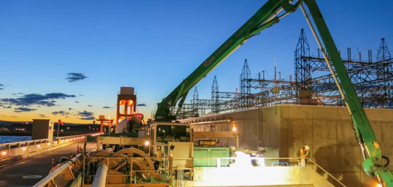 Concrete pump truck with a long green boom works at an industrial site by a waterway at dusk, with workers in safety gear and power grid structures silhouetted against the sunset.