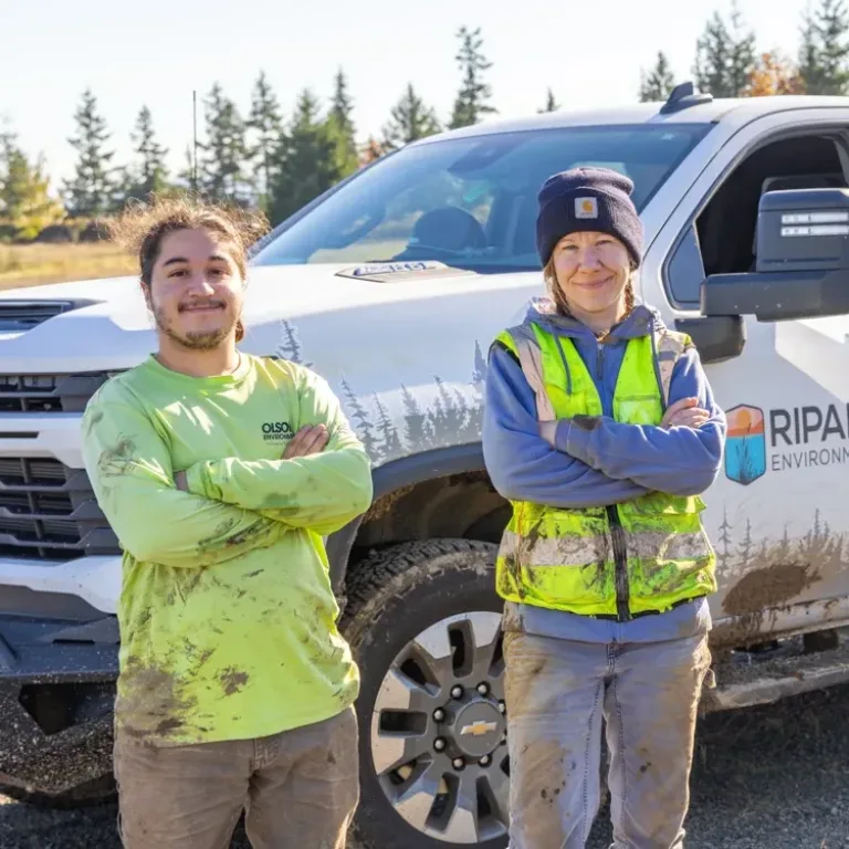 MacKay field crew members stand with arms crossed beside a mud-splattered pickup truck, wearing high-visibility safety gear at an outdoor job site.