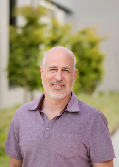 Headshot of Rob Palena, a smiling man with a shaved head and short gray beard wearing a light purple polo, outdoors with blurred greenery in the background.