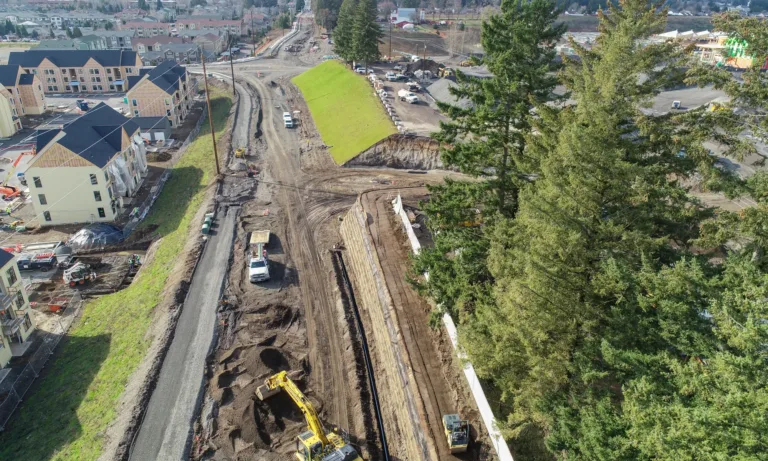 Aerial view of a suburban road construction site with excavators and trucks shaping a gravel roadbed and retaining wall between new apartment buildings and tall evergreen trees.