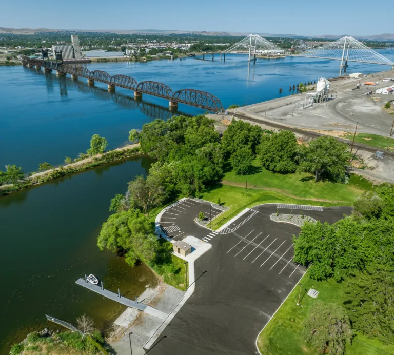 Aerial view of a riverside park and boat launch with an empty parking lot and dock beside a narrow lagoon, opening to a wide river spanned by an old arched truss railroad bridge and a modern cable-stayed bridge on a clear day.