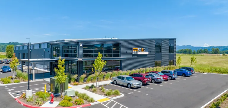 Modern two-story commercial building with dark gray metal siding and large glass windows, a landscaped parking lot with cars, and open fields with mountains in the background under a clear blue sky.