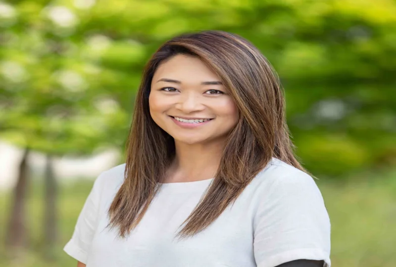 Headshot of Stephanie Zhao, smiling outdoors with straight brown hair and a light gray top against a blurred green background.