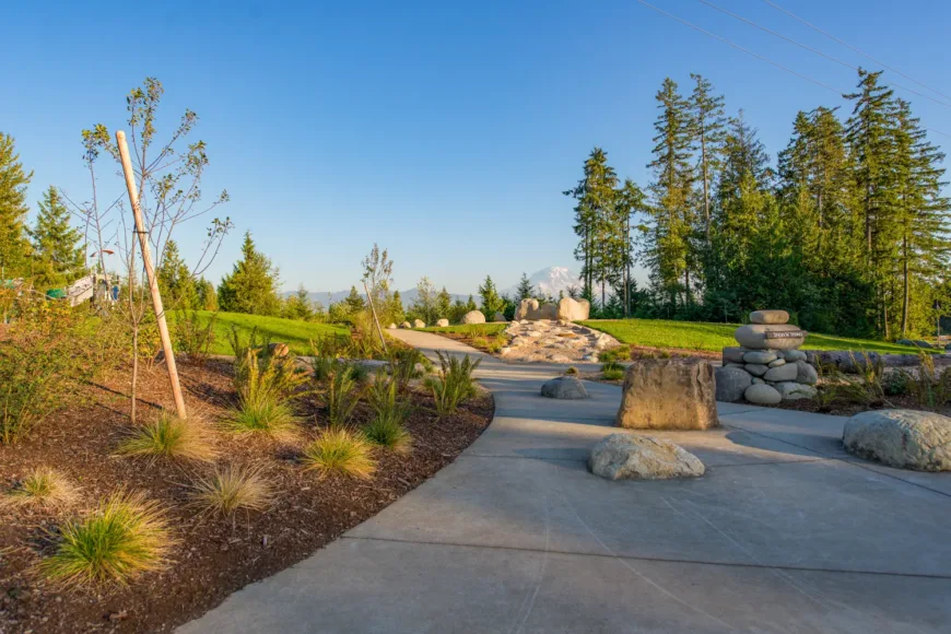 Curving paved pathway through a landscaped park with boulders, native grasses, and evergreen trees, featuring a stacked cairn labeled Story Stone and a distant snow-capped mountain under a clear blue sky.