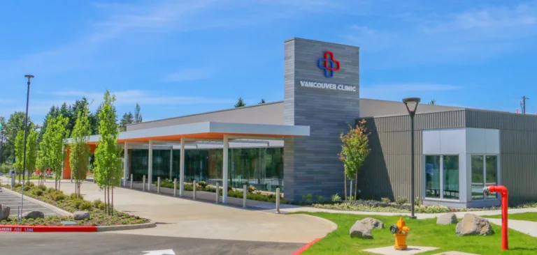 Modern single-story medical clinic with a glass-front entrance and covered drop-off, a sign tower reading “VANCOUVER CLINIC,” landscaped trees, and a sunny blue sky.