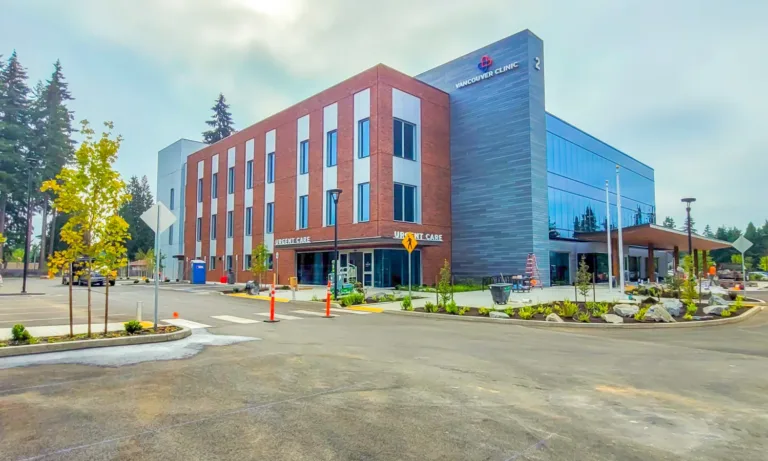 The Vancouver Clinic building with an Urgent Care entrance, featuring a brick-and-glass facade and a mostly empty parking lot with young trees on an overcast day.