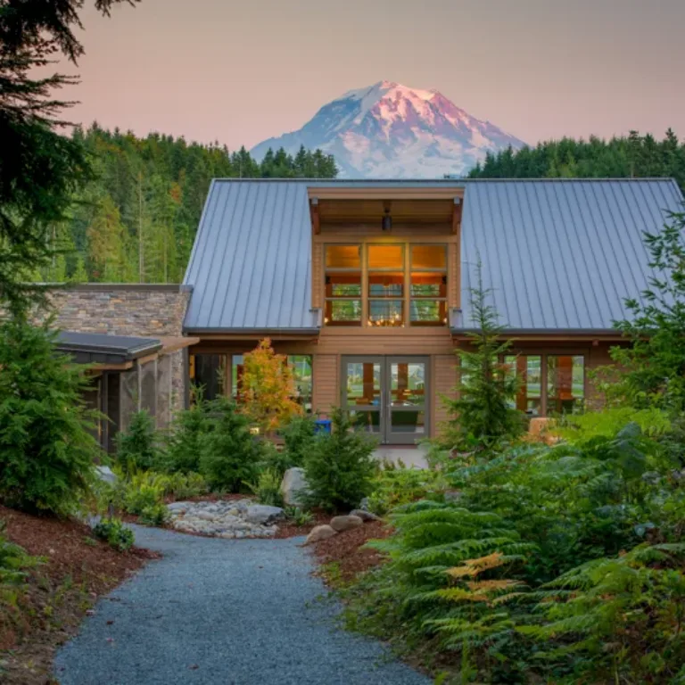 A modern timber house with a steep metal roof and large windows sits amid lush evergreens, a gravel path leading to the entrance, with a snow-capped mountain glowing at sunset in the background.