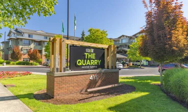 Entrance sign for Cogir The Quarry Senior Living at 415, with landscaped lawns, flowers, and the multi‑story community buildings in the background on a sunny day.