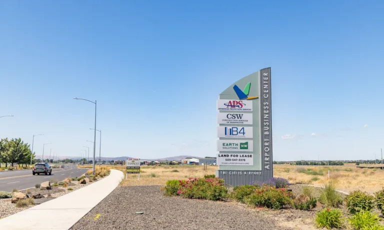 Roadside monument sign for the Airport Business Center near Tri-Cities Airport listing APS Advanced Protection Services, CSW Construction Services of Washington, IB4, Earth Solutions, and Land for Lease, set against an open landscape and clear blue sky.