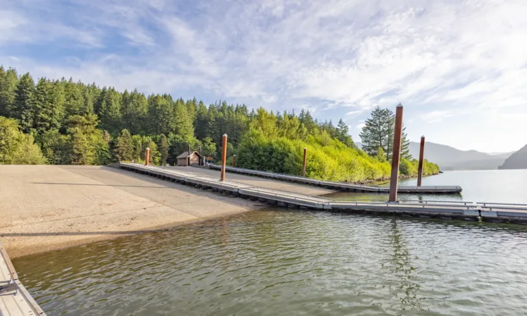 Boat launch with floating docks and tall pilings on a calm lake, backed by dense forested hills and a small building under a partly cloudy sky.