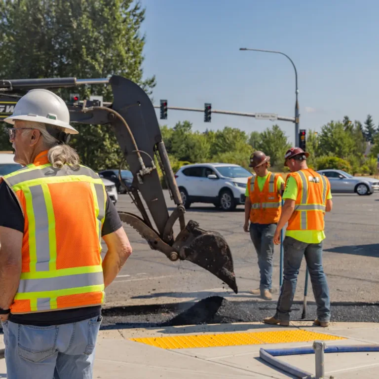 Road crew in orange safety vests and hard hats working by an excavator at a sunny intersection with fresh asphalt and cones.