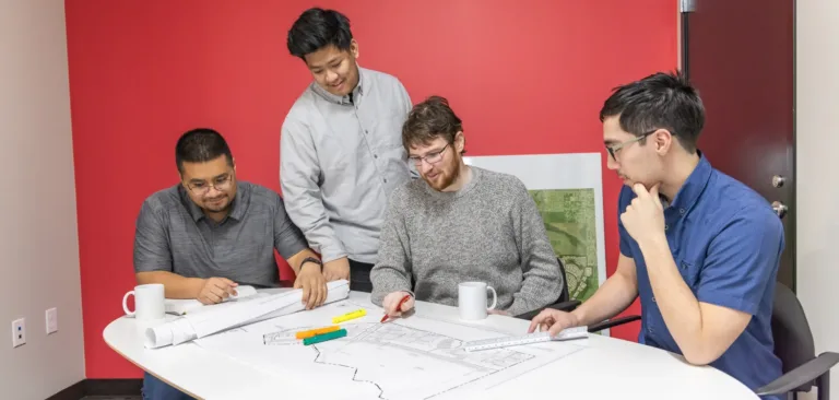 Four colleagues review architectural plans at a white table, discussing details with pens and mugs in a modern red-walled office.