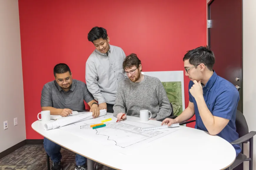 Four colleagues review architectural plans at a white table, discussing details with pens and mugs in a modern red-walled office.