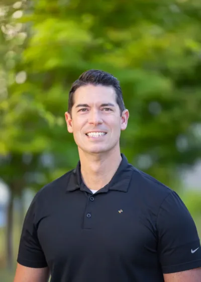 Smiling man in a black polo shirt standing outdoors with green foliage in the background