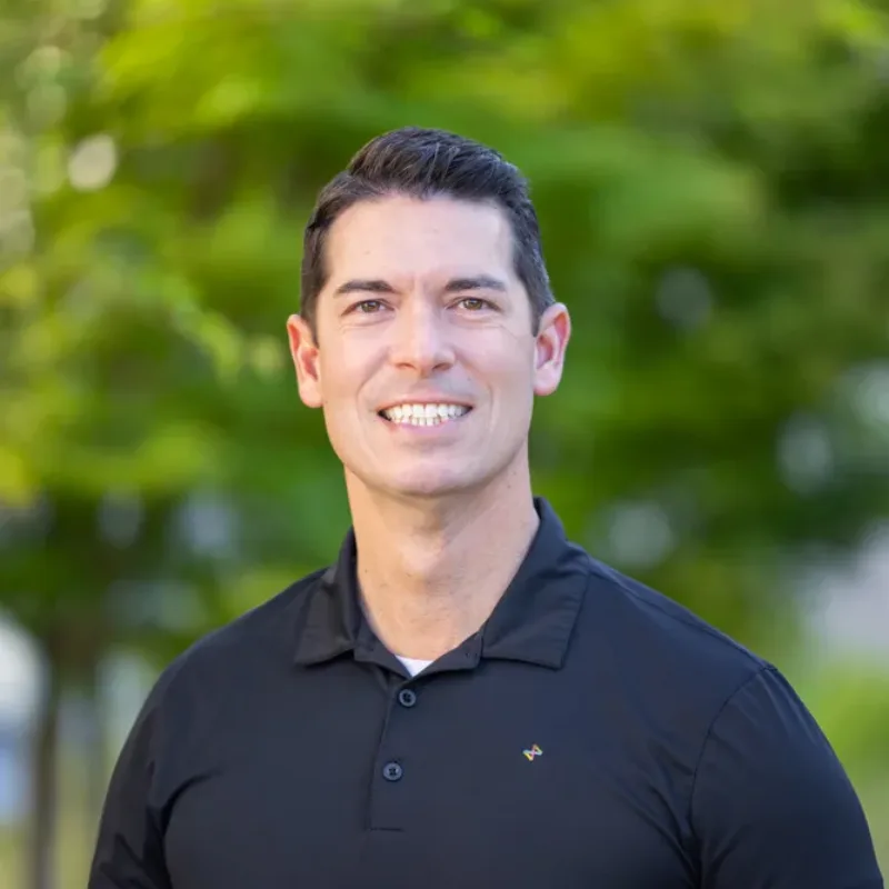 Smiling man in a black polo shirt standing outdoors with green foliage in the background