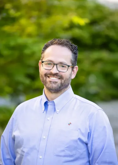 Man with glasses and a short beard smiling in a light blue button-down shirt, standing outdoors with green foliage behind him