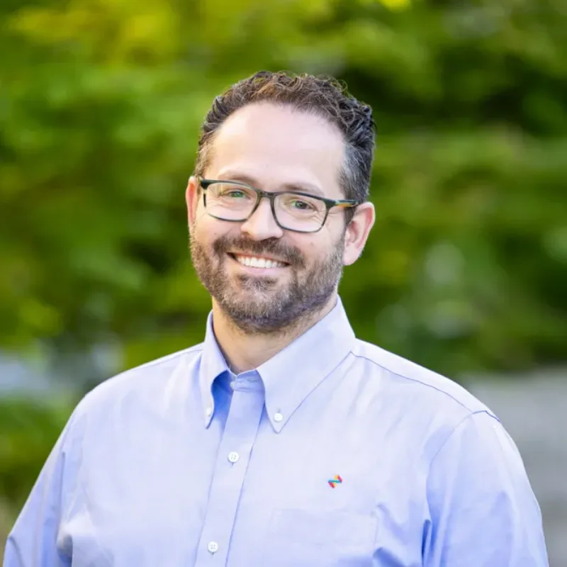 Man with glasses and a short beard smiling in a light blue button-down shirt, standing outdoors with green foliage behind him