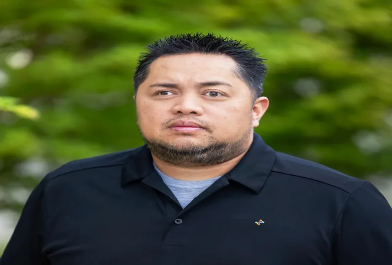 Man with short spiked hair and a trimmed beard wearing a black polo, standing outdoors with blurred green foliage behind him