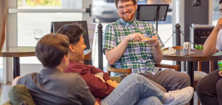 MacKay team members relax and chat in a casual office lounge during a social, with one person smiling and holding a plate of snacks.