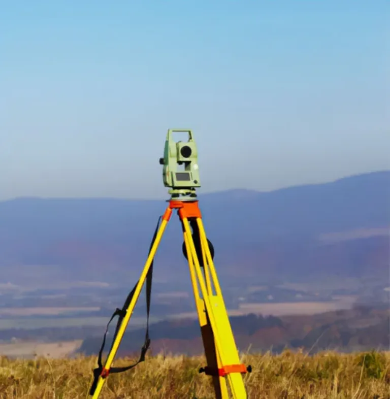 Green surveying total station on a yellow tripod in a grassy field with distant hills under a clear blue sky.