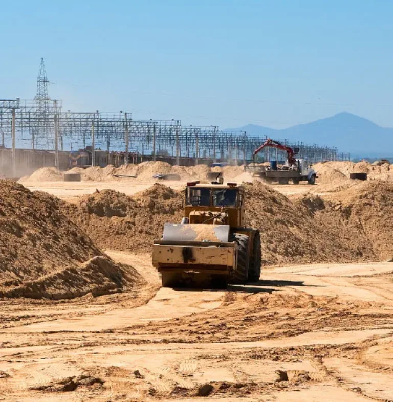 Bulldozer grading a sandy construction site beside power lines, with an excavator working in the background and distant mountains under a clear sky.