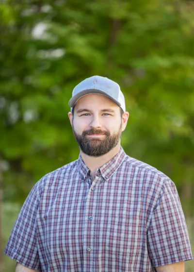 Bearded man in a gray cap and plaid shirt smiling outdoors with a green, blurred background