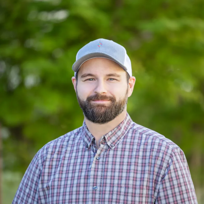 Bearded man in a gray cap and plaid shirt smiling outdoors with a green, blurred background