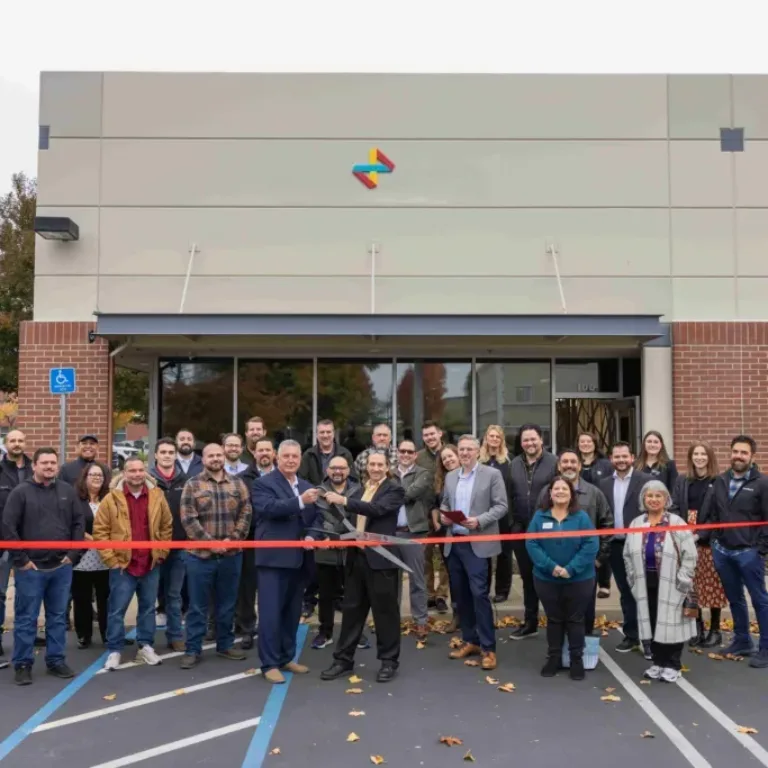 Group of people at a ribbon-cutting ceremony in front of a building, with a red ribbon stretched across the parking lot.