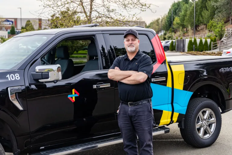 Man in a black polo and cap stands with arms crossed beside a black pickup truck with colorful geometric decals.