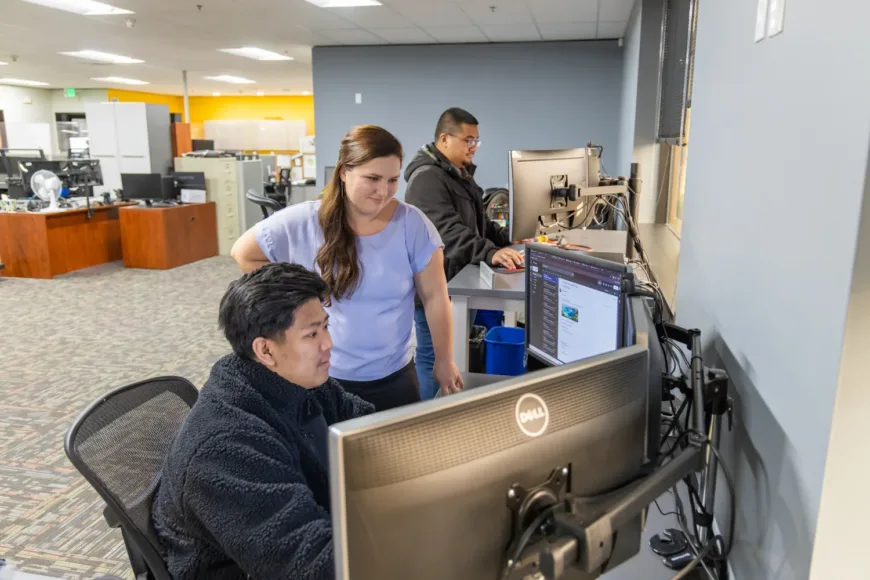 Three coworkers collaborate at computer workstations in a modern office, with one seated and two standing, focused on screens.