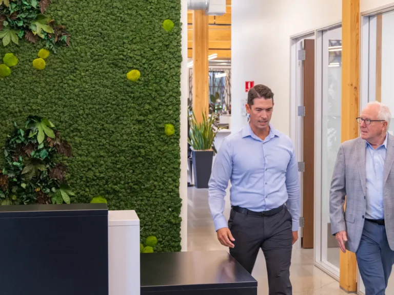 Two men in business attire walk through a modern office corridor with a green moss wall and glass-walled meeting rooms.