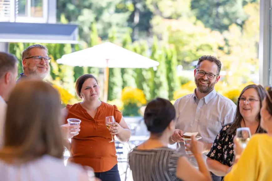 Group of adults chatting and laughing at an outdoor gathering, holding drinks, with sunny greenery and umbrellas in the background.
