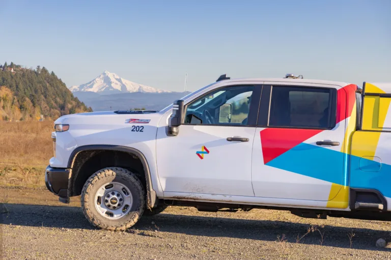 White utility pickup with colorful geometric decals parked on a gravel road, snow-capped mountain in the background under clear sky