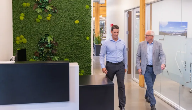 Two men in business attire walk through a modern office hallway with a green moss wall and glass-walled meeting rooms.