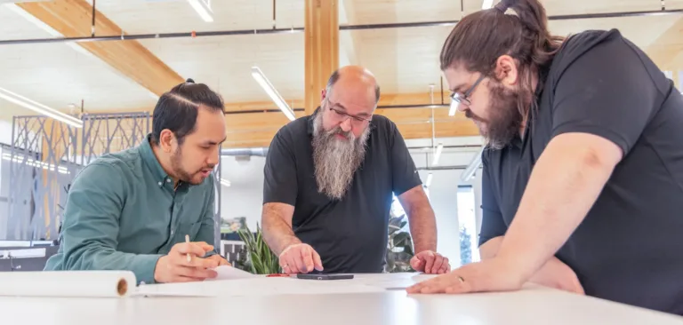 Three coworkers in a modern office lean over a table, discussing documents and sketches together.