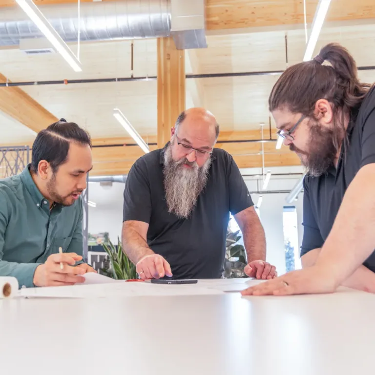 Three coworkers in a modern office lean over a table, discussing documents and sketches together.