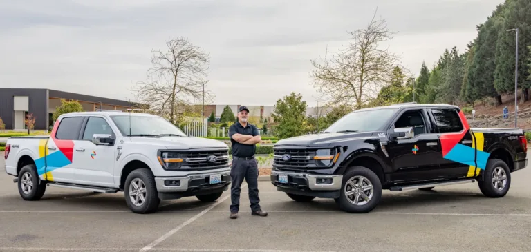 Two branded pickup trucks, one white and one black, parked side by side with a uniformed worker standing between them.